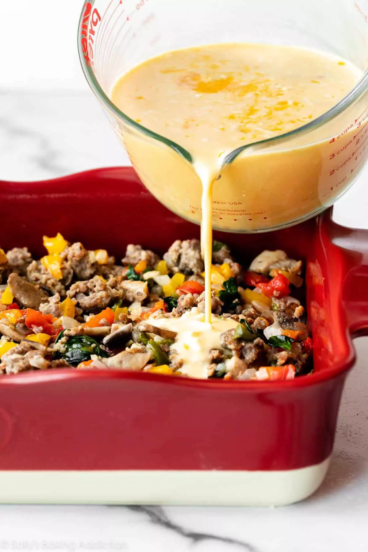pouring egg mixture over sausage, vegetables, and bread in red casserole dish.