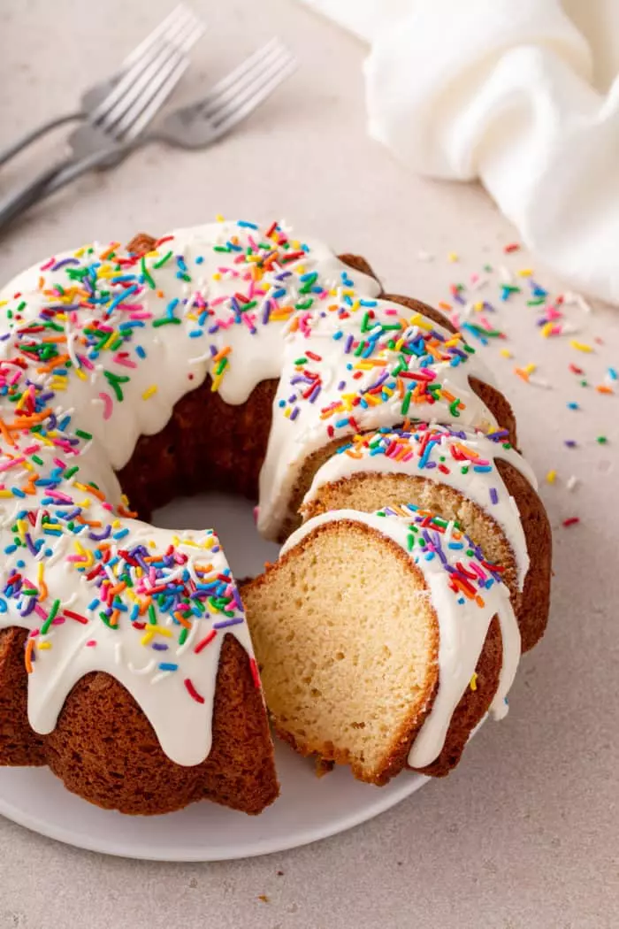 Two slices of vanilla bundt cake topped with cream cheese frosting and rainbow sprinkles laying down on a white plate.