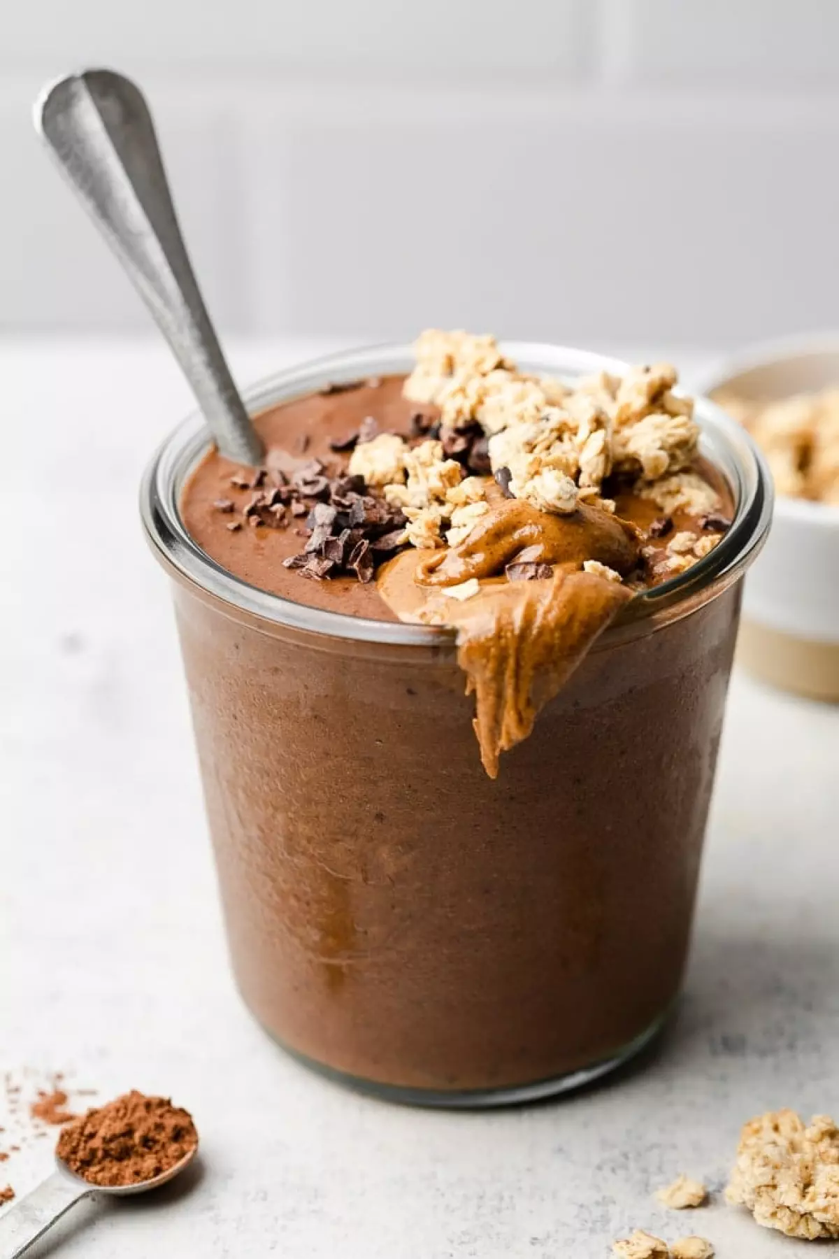 Chocolate protein smoothie in a glass jar topped with granola and nut butter. There is a spoon in the jar as well.
